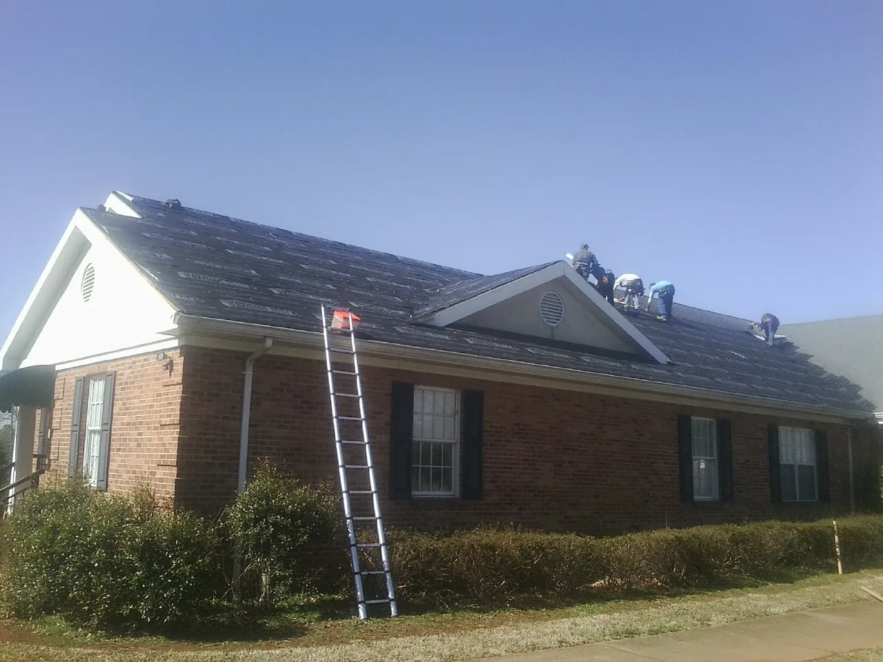 Crew tearing shingles off a brick ranch roof with a ladder set — Metal Roof Repair work by our crew in Logandale