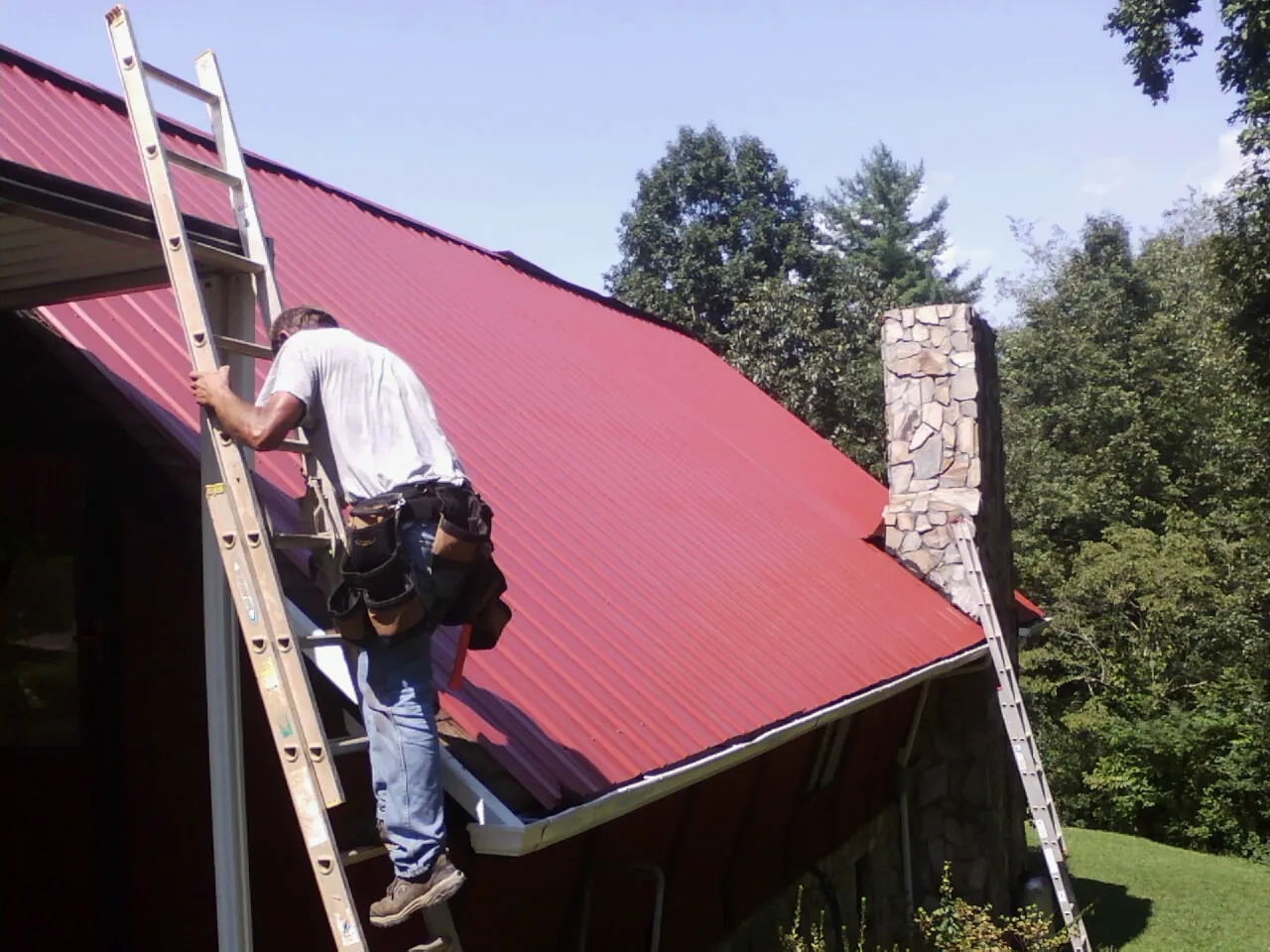 Roofer on a ladder beside a red metal roof and stone chimney — Soffit & Fascia Repair by Roofing Brothers in Logandale
